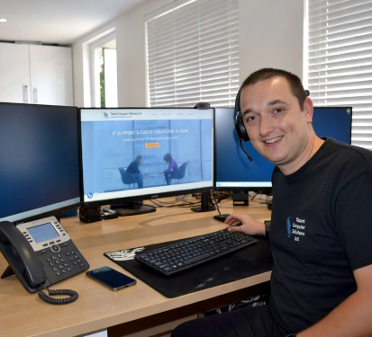 Smiling IT support technician at his desk, wearing a headset with multiple monitors displaying an IT support website.